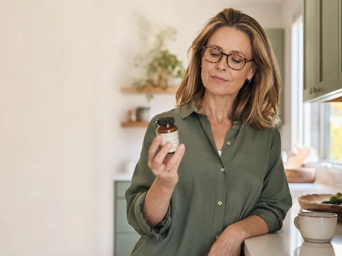 A woman in her early fifties calmly reading the label on a supplement bottle in a bright kitchen, looking thoughtful and discerning.